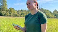 Nainen seisoo vihreällä niityllä pitäen kädessään älypuhelinta, taustalla metsää ja sininen taivas.
