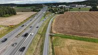Useita autoja liikkuu monikaistaisella tiellä maaseutumaisessa maisemassa, taustalla metsää ja rakennuksia.