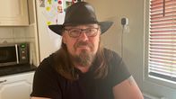 Pitkähiuksinen, silmälasipäinen mies, jolla on musta hattu ja t-paita, istuu keittiössä pöydän ääressä.