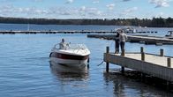 Mies moottoriveneessä Vääksyn Majakkapaviljongin satama-alueella kiinnittymässä laituriin. Laiturilla mies ja nainen pitävät kiinni veneen köysistä.