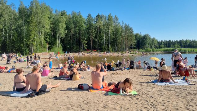 Many people on a sandy beach, with green trees and a lake in the background.