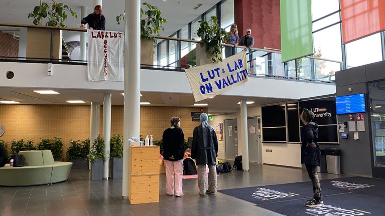 Kuusi opiskelijaa aulatilassa virittämässä lakanoita, joissa mielenosoitustekstejä.