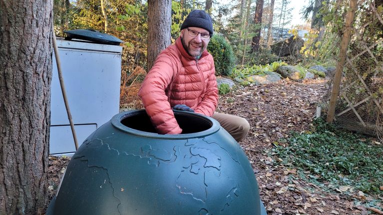 Puutarhuri Marko Ahola käsi maapallokompostorissa, takana isoja mäntyjä ja pakastimesta rakennettu kompostori