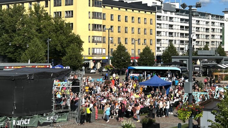 Lähikuvassa on aukea tori, jossa on ihmisiä ja musta esiintymislava. Ympärillä on rakennuksia. 