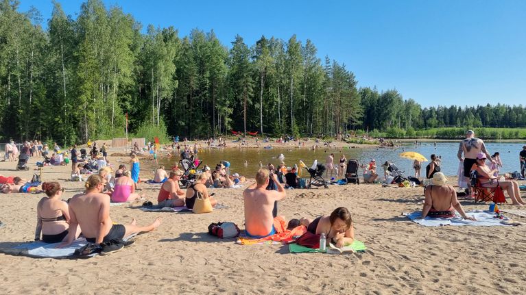 Many people on a sandy beach, with green trees and a lake in the background.