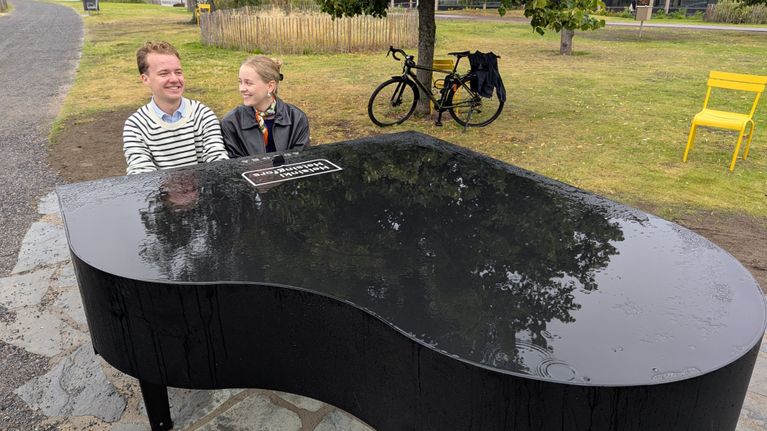 Puistossa on iso, kastunut puistoflyygeli, jonka edessä istuvat mies ja nainen soittamassa.