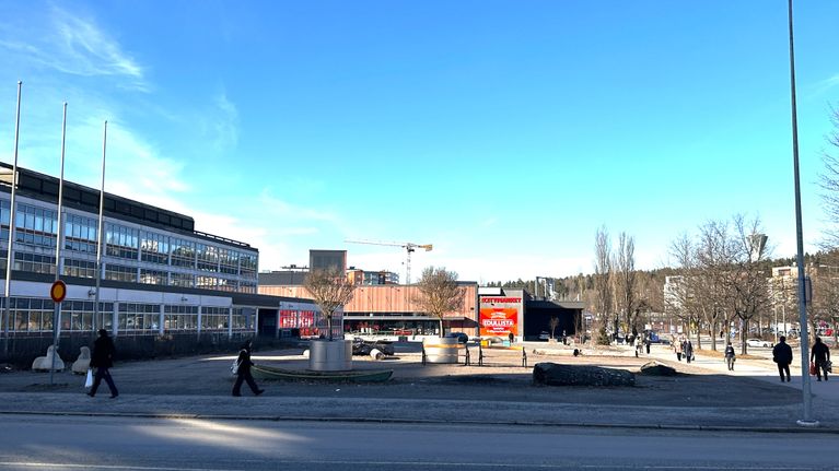 Kuvassa on hiekkainen maatontii, jonka ympärillä on rakennuksia ja kävelyteitä. Teillä on ihmisiä. 