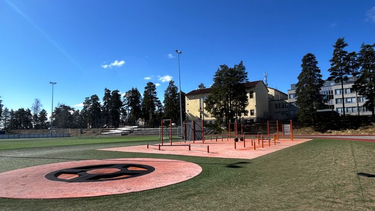 Kuvassa on urheilukenttä, jossa on punapohjainen urheilualue. Taustalla on keltainen suuri rakennus. 
