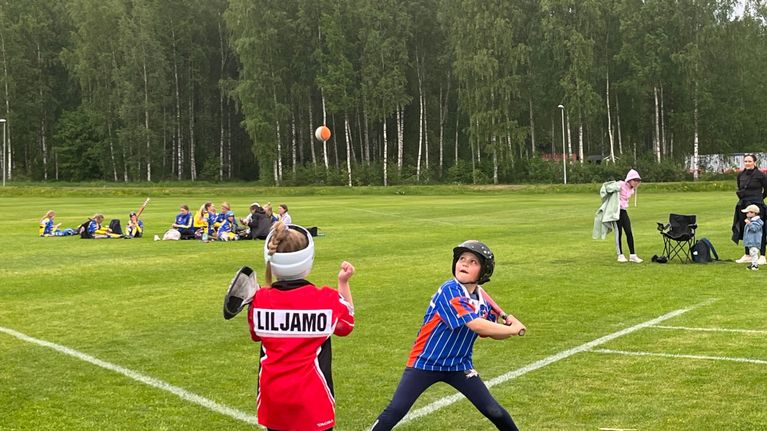 En spelare i blå tröja svingar ett slagträ mot en boll medan en spelare i röd tröja utövar försvar på en gräsplan, med fler personer i bakgrunden.