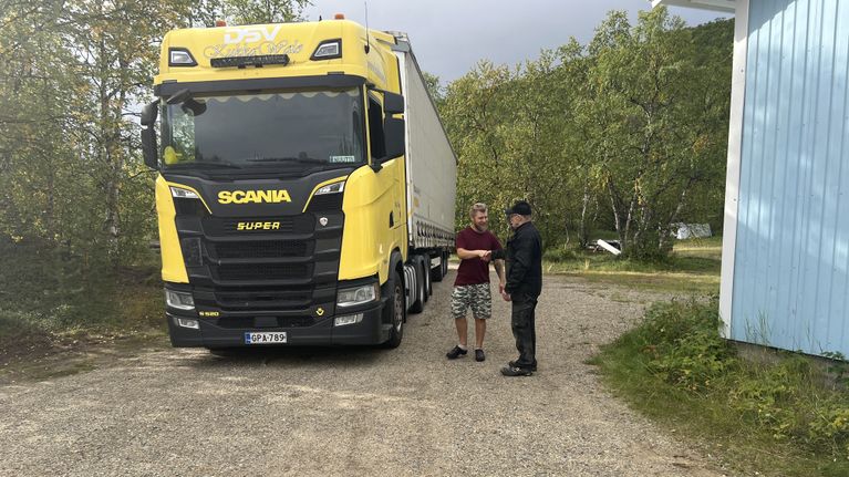 Kaksi henkilöä seisoo hiekkatiellä suuren keltaisen Scania-rekan edessä rakennuksen vieressä. Rekassa tekstiä ”DSV” ja ”Scania Super”.