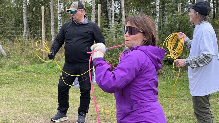 Kolme aikuista seisoo nurmikolla, taustalla metsää. Porohoitajat harjoittelevat suoppunginheittoa varten, 29.8.2026 Reindeer Manor Levillä. Major.