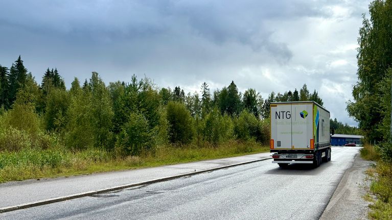 Valkoinen rekka ajaa tiellä. Tien vieressä on tyhjä tontti, jossa kasvaa lehtipuita.