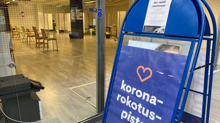 Koronarokotuspiste -teksti a-standissä, jonka takana näkyy tyhjä tila, jossa on tuoleja.