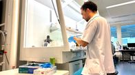 Valkoiseen tutkimustakkiin ja sinisiin kertakäyttökäsineisiin pukeutunut ruskeahiuksinen mies katselee näytettä tutkimuspöydän ääressä laboratoriossa.