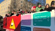 Naamiaisasuisia nuoria ihmisiä kuorma-auton lavalla. Lavan reunasta roikkuu julisteita. Toisessa on punareunainen kolmio, jonka sisällä on ylioppilaslakki. Alla lukee varokaa juhlivia abeja. 