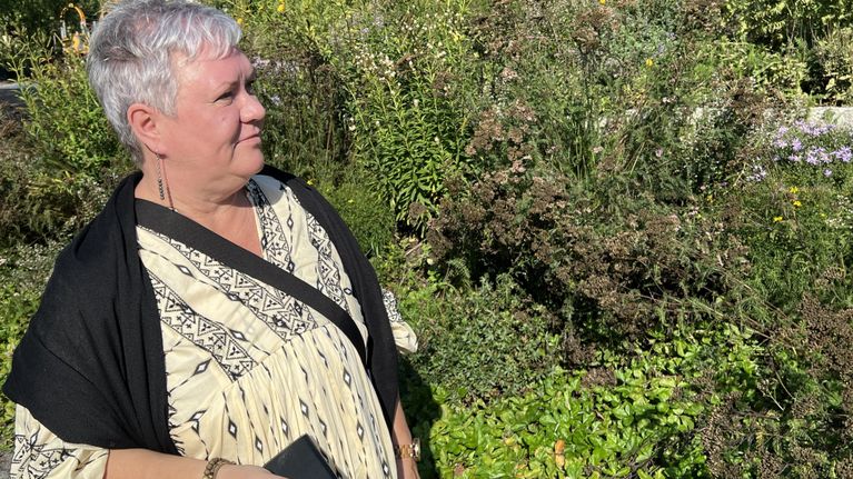 En kvinna med kort grått hår och mönstrad klänning står utomhus vid en grönskande rabatt och håller en mobiltelefon i handen.