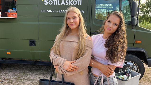 Two young women stand in front of a green army van.