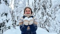 Silmälasipäinen nainen villapaita päällään seisoo lumen peittämien kuusien keskellä ja kannattelee käsissään rukkasten päällä pientä kasaa lunta.