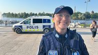 Silmälasipäinen iloisesti naurava poliisinainen lippalakki päässä, taustalla jokirantaan parkeerattu poliisiauto.