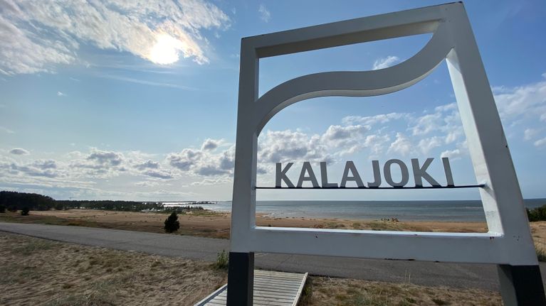 Puinen kyltti, jossa lukee Kalajoki. Taustalla merenrantaa.