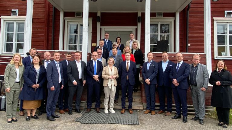 Ryhmäkuva, jossa joukko ihmisiä seisoo hirsitalon edustalla. 