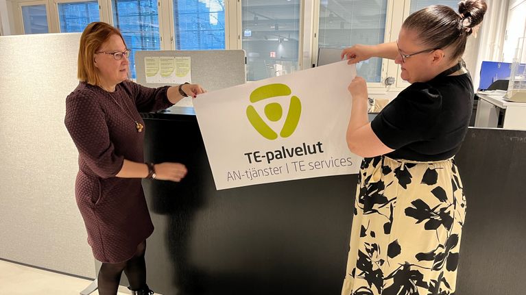 Kaksi naista pitelee tiskin vieressä julistetta, jossa lukee TE-palvelut. 