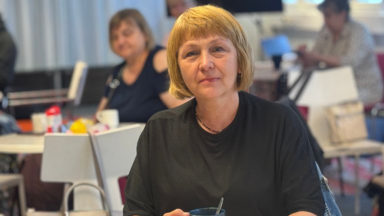 A woman with short blonde hair sits at a table holding a dark mug, with other people seated in the background.