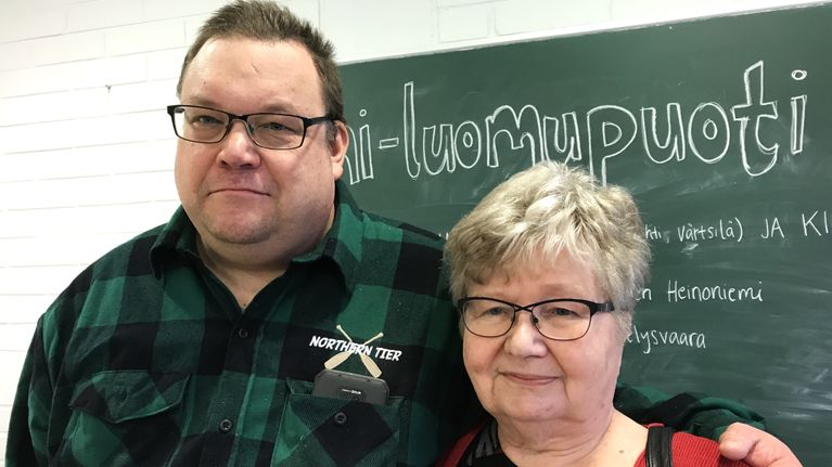 Silmälasipäiset mies ja nainen seisovat vierekkäin koulun liitutaulun edessä.