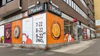 Rakennuksen kulmassa on K-Market. Ikkunoissa on kaupan mainoksia ja aukioloajat. 