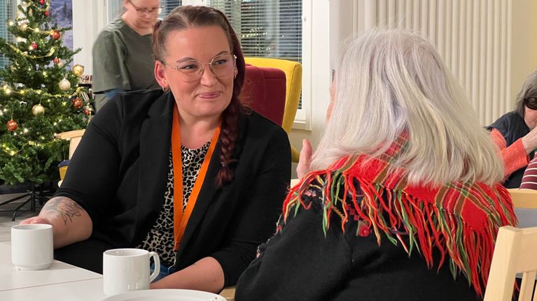 Maria Jaakkola istuu kahvipöydässä ja jutustelee palvelutalon asukkaan kanssa.