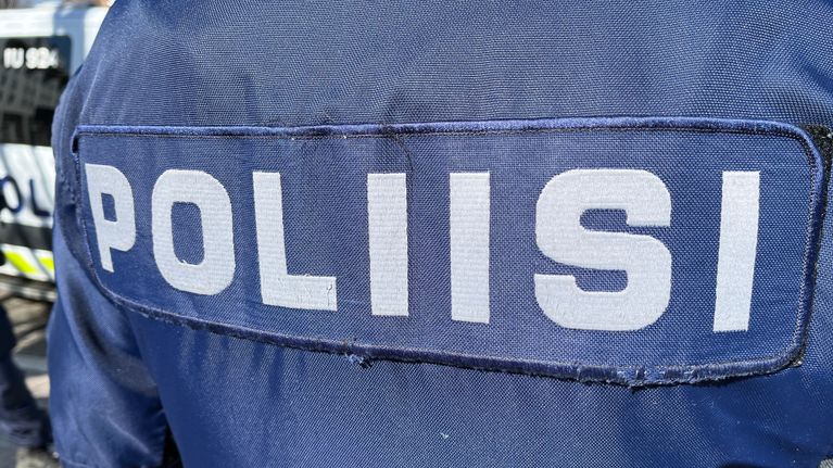 Sinisen haalarin selkä, jossa lukee "poliisi". Taustalla näkyy hieman poliisiautoa.