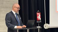 Mies puvussa puhuu salin edessä. Vieressä on Orimattilan punainen viiri, jossa on kuva valkoisesta hevosesta.