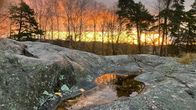 Puiden takana loistava auringonnousu heijastuu pienestä lammikosta kalliolla.