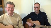 Två män sitter med gitarrer i famnen och tittar in i kameran. 