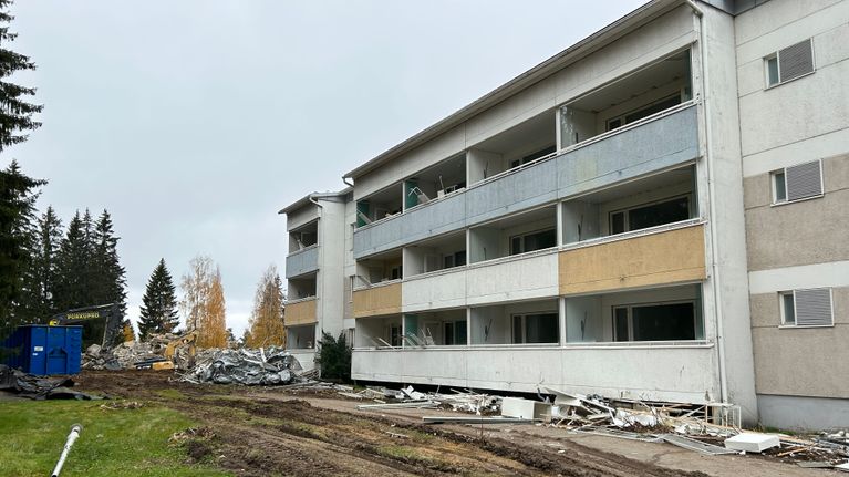 Purettava kerrostalo, purkujätettä maassa ja työkoneet käsittelemässä purkujätettä taustalla.