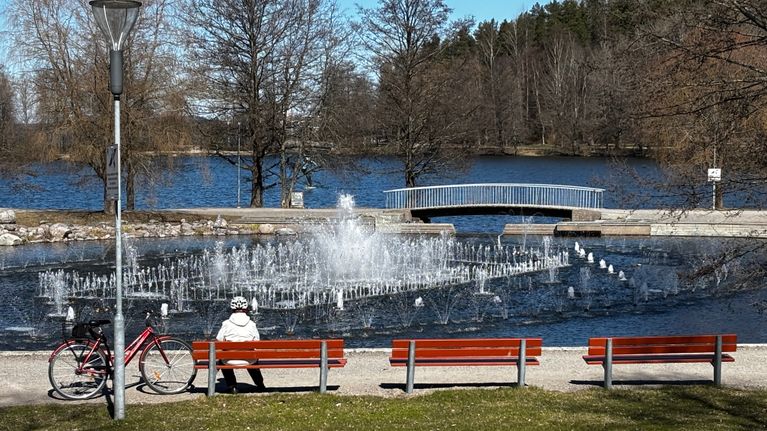 Puiston penkillä istuu ihminen pyörä vieressään. Hän katsoo kohti suihkulähdettä ja järveä. Etualalla näkyy nurmikkoa. Puissa ei vielä ole lehtiä.