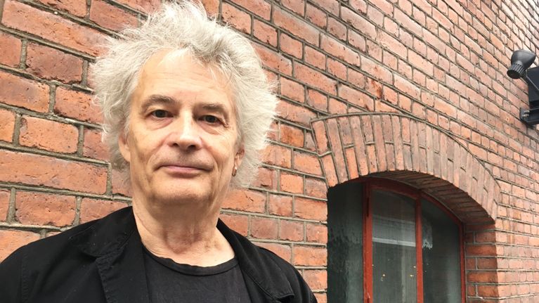 Harmaatukkainen mies seisoo kuvan vasemmassa laidassa, taustalla tiiliseinä. Kuvassa on Eppu Normaali-yhtyeen Juha Torvinen.