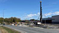 Rakennustyömaa tien varressa, suuri paalutuskone, työajoneuvoja ja hallirakennus oikealla, taustalla metsää.