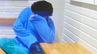 En person i blåa kläder sitter vid ett träbord i ett ljust rum och håller ena armen framför ansiktet.
