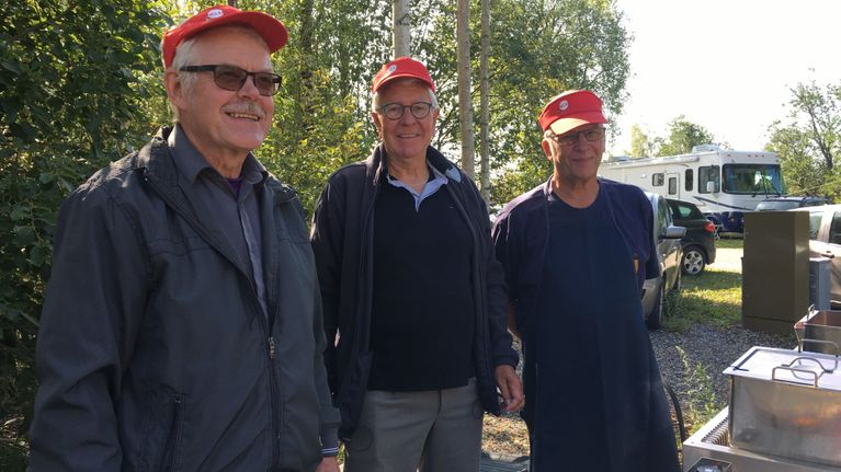 Tre män med röda skärmmössor står vid en korvgrill.