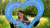 Anne-Marie Kalla pitelee minä rakastan Inaria -kylttiä, taustalla joki ja vihreää kasvustoa. 