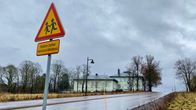 En gammal grågrön stenbyggnad i två våningar, med en flygel bredvid i en våning. I förbrunden ett trafikmärke som varnar för barn. Våt asfaltväg. Lövträd som fällt sina löv. Höststämning. 