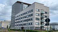 Vaaleanharmaa, useampikerroksinen asuinkerrostalo pilvisenä päivänä, etualalla tie ja villiintynyttä kasvillisuutta.