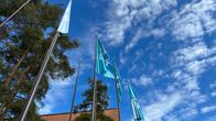 Puolipilvinen taivas, edustalla mäntyjä ja lipputankoja, joissa turkoosit liput. Puiden takana punainen tiilirakennus.