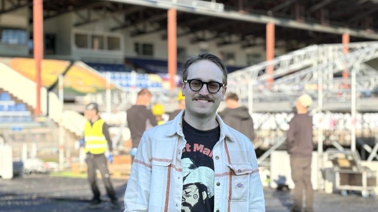 En man med glasögon och mustasch står utomhus framför en scenkonstruktion, Andra personer och läktare syns i bakgrunden.