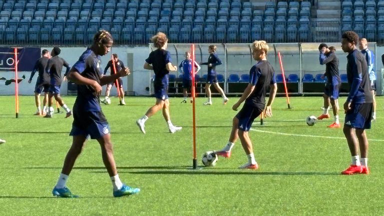 Flera fotbollsspelare tränar på en gräsplan med koner och bollar, tomma läktare i bakgrunden.