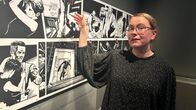 Nainen osoittaa seinällä olevaa mustavalkoista sarjakuvataulua.