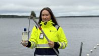 En person i gul varseljacka håller en vattenprovtagare och en flaska vid en brygga intill en sjö. 