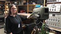 Silmälasipäinen nainen seisoo teknisiä laitteita sisältävien hyllyjen edessä ja pitää hansikoidulla kädellä kiinni vanhasta jaloilla seisovasta tv-kamerasta, jossa lukee Suomen Televisio. 