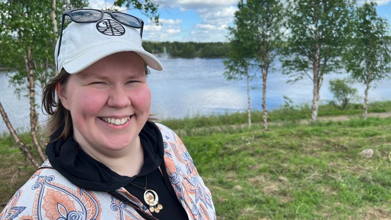 Kuvassa ihminen hymyilee leveästi veden äärellä. Päässään hänellä on lippalakki ja otsalla aurinkolasit. 